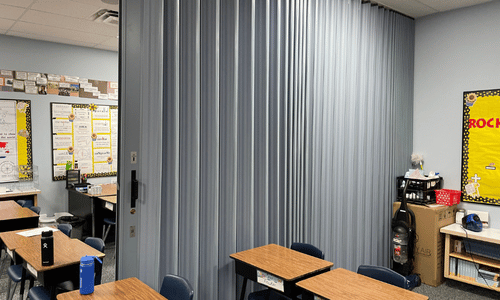 A folding divider partially separates a classroom with desks, chairs, and educational posters on the walls, near the commercial doors leading to the hallway.