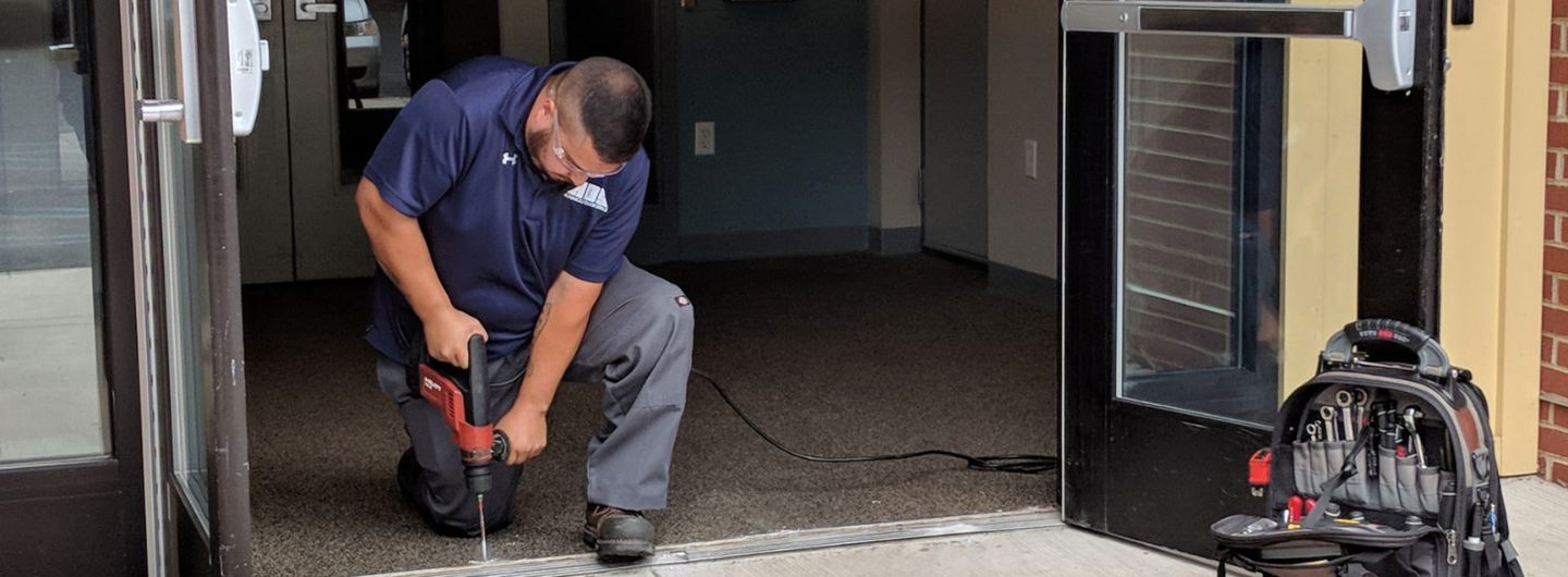 A person kneels while using a drill on the floor near an open doorway, working on commercial door repair, with a tool bag on the right.