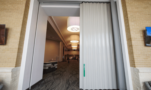 Large accordion-style commercial door partially opened, revealing a spacious, modern interior with pendant lights and patterned carpeting—ideal for businesses seeking reliable commercial door installation.